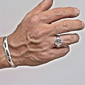 May include: A silver cuff bracelet and a silver signet ring. The cuff bracelet has a textured design with dark accents. The ring features a detailed emblem on its face. Both pieces are displayed on a hand against a neutral background.