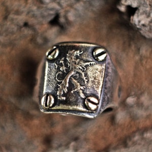 May include: A silver ring with a brass lion crest design. The ring has four gold screws on the sides.