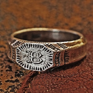 Könnte beinhalten: Ein silberner Ring mit einem achteckigen Design, das einen erhabenen Buchstaben "B" in der Mitte zeigt. Der Ring hat eine strukturierte, abgenutzte Optik, was darauf hindeutet, dass er antik ist.