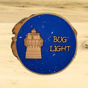 May include: A wooden round sign with a blue background and white speckles. The sign features a laser-cut image of a lighthouse with the text "BUG LIGHT" in brown letters.