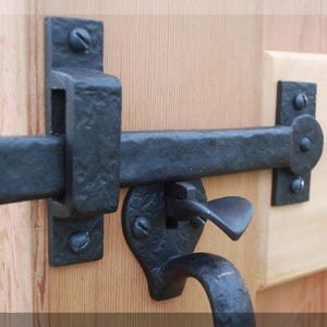 May include: Close-up of a black, wrought iron gate latch. The latch features a handle, a sliding bar, and a mounting plate, all attached to a light-coloured wooden surface. The hardware is secured with black screws.