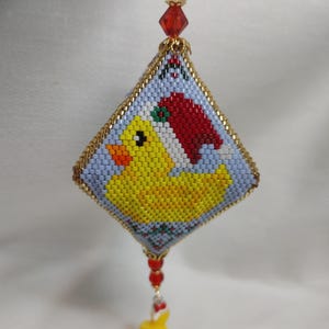 May include: A beaded Christmas ornament in a diamond shape, featuring a yellow duck wearing a red Santa hat. The ornament has a light blue background and gold trim. A yellow pom-pom hangs from the bottom.