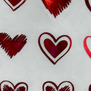 May include: A sheet of red heart stickers with a white outline. The hearts have a shiny, metallic finish and are decorated with a hatched pattern.