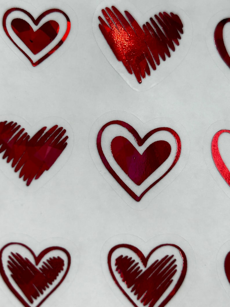 May include: A sheet of red heart stickers with a white outline. The hearts have a shiny, metallic finish and are decorated with a hatched pattern.