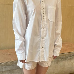 Op de afbeelding: Een wit overhemd met lange mouwen, knopen, een staande kraag en een rij zilveren knopen aan de voorkant. Het overhemd wordt gedragen over een wit kanten broekje.