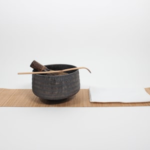 May include: A Japanese tea ceremony set, comprising a dark grey ceramic bowl, bamboo whisk, and scoop, on a bamboo mat. A white cloth is placed to the right. The items are set against a white background.