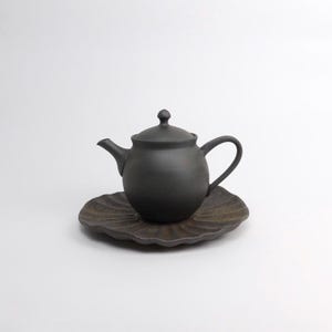 Puede incluir: Una tetera de cer&aacute;mica negra mate con tapa y asa a juego se asienta sobre un platillo marr&oacute;n oscuro en forma de hoja. La tetera tiene un cuerpo redondeado y un pico curvo. El juego est&aacute; dise&ntilde;ado para servir t&eacute;.