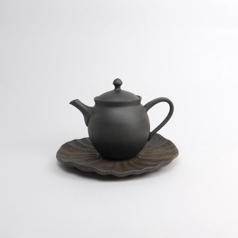 Puede incluir: Una tetera de cer&aacute;mica negra mate con tapa y asa a juego se asienta sobre un platillo marr&oacute;n oscuro en forma de hoja. La tetera tiene un cuerpo redondeado y un pico curvo. El juego est&aacute; dise&ntilde;ado para servir t&eacute;.