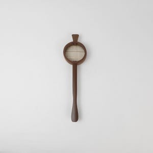 May include: A brown wooden tea strainer with a circular head and a long handle. The strainer features a woven mesh screen within the circular frame. The handle is smooth and tapers towards the end, creating a simple, elegant design.