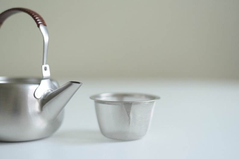 Może przedstawiać: Srebrny czajnik z brązowym sk&oacute;rzanym uchwytem i pasującym srebrnym sitkiem do herbaty.