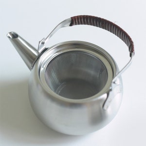 Może przedstawiać: Srebrny czajnik z wbudowanym sitkiem i brązowym sk&oacute;rzanym uchwytem.