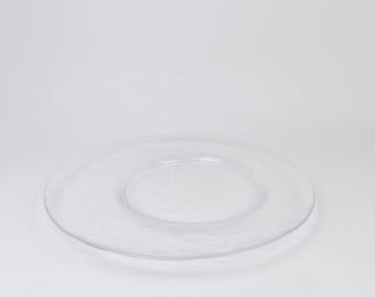 May include: A clear glass plate with a textured surface. The plate is round with a slightly raised rim and a shallow centre. The plate is set against a white background.