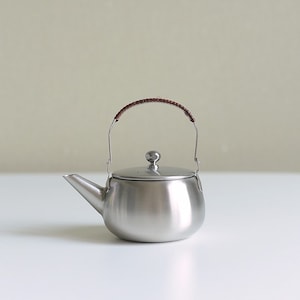 Może przedstawiać: Srebrny czajnik ze stali nierdzewnej z brązowym sk&oacute;rzanym uchwytem. Czajnik ma okrągły korpus i pokrywkę z małym guzikiem na g&oacute;rze.
