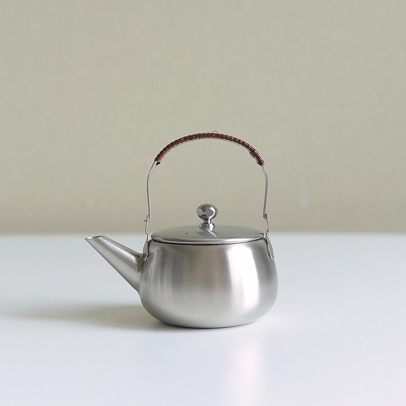 Może przedstawiać: Srebrny czajnik ze stali nierdzewnej z brązowym sk&oacute;rzanym uchwytem. Czajnik ma okrągły korpus i pokrywkę z małym guzikiem na g&oacute;rze.