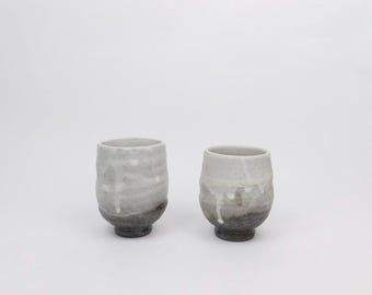 May include: Two handmade ceramic cups. The cups are cylindrical with a slightly flared rim and a dark base. The glaze is a mix of white and gray, with drips of glaze adding texture. The cups are approximately 3 inches tall.