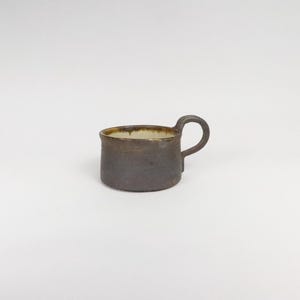 Peut inclure: Une petite tasse en céramique brun foncé avec un bord brun clair et une anse. La tasse est posée sur une surface blanche.