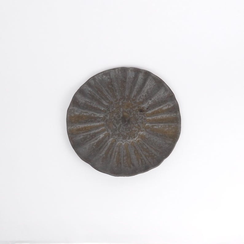 Puede incluir: Un plato decorativo redondo, de color bronce oscuro, con un dise&ntilde;o texturizado y acanalado que irradia desde el centro. El plato tiene un borde ligeramente irregular y festoneado. El plato est&aacute; sobre un fondo blanco liso.