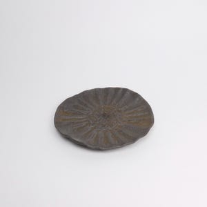 Puede incluir: Un plato de cer&aacute;mica gris oscuro con una superficie texturizada y un dise&ntilde;o de rayos de sol. El plato tiene una forma org&aacute;nica e irregular y probablemente est&aacute; hecho a mano. Es un art&iacute;culo decorativo.