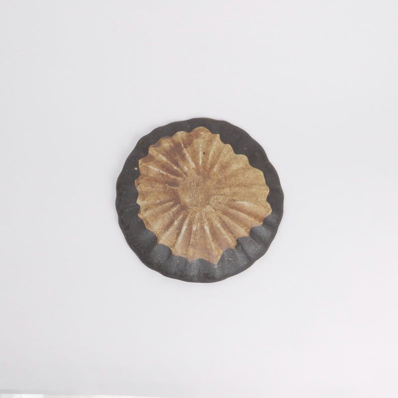 Puede incluir: Un plato redondo de cer&aacute;mica con un borde marr&oacute;n oscuro y un centro beige texturizado. El centro tiene un patr&oacute;n radial y un peque&ntilde;o dise&ntilde;o en relieve. El plato est&aacute; sobre un fondo blanco.