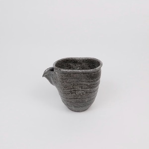 May include: A dark grey ceramic pitcher with a spout. The handmade pitcher has a textured surface and an organic shape. The pottery is likely stoneware, suitable for serving liquids.