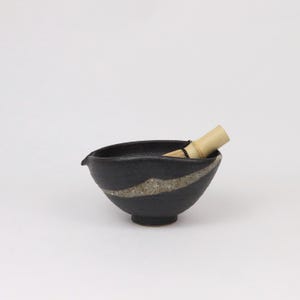 Puede incluir: Un cuenco de matcha de cerámica negra con una franja de color claro y un batidor de bambú. El cuenco tiene un pequeño pico y una base redondeada. El batidor es de bambú y está parcialmente dentro del cuenco.
