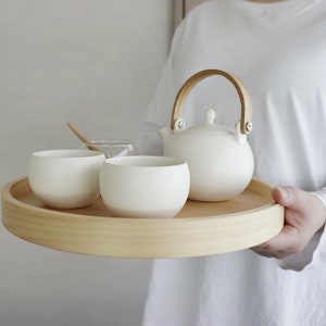 Może przedstawiać: Biały ceramiczny czajnik z drewnianym uchwytem, dwie białe ceramiczne filiżanki i drewniana taca. Czajnik i filiżanki znajdują się na tacy.
