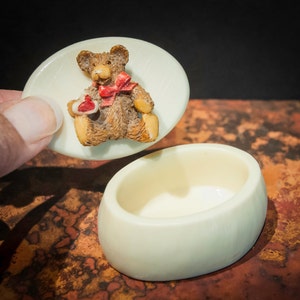 May include: A small white ceramic trinket box with a lid featuring a brown teddy bear figurine holding a red heart. The bear has a red bow around its neck.