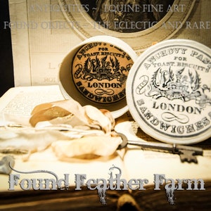 May include: Two vintage white ceramic jars with black and white labels. The labels read "Anchovy Paste for Toast Biscuits London Sandwiches &" and "Anchovy for Toast Biscuits London Sandwiches". The jars are sitting on a book with the words "Found Feather Farm" printed on the cover.