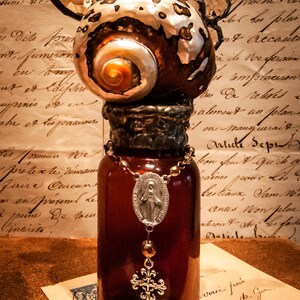 May include: A glass bottle with a brown liquid inside. The bottle is decorated with a gold cross and a silver charm with a religious image. The bottle is topped with a decorative shell with a silver and gold finish. The bottle is sitting on a brown surface with a piece of paper that says "Found Feather Farm".