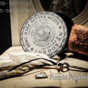 May include: A vintage, round, white tin container with black lettering that reads "Home Made Potted Meats Manufactured by the Army and Navy Co-Operative Society 105 Victoria St. London S.W. 427". The tin is sitting on a book with a feather and a key.