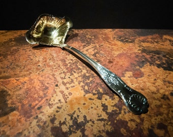 May include: A silver ladle with an ornate handle and a decorative, scalloped bowl. The ladle is lying on a rustic, brown surface.