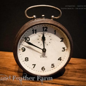 May include: A vintage, round, black alarm clock with white face and black hands. The clock face has black numbers and a small black 'Bravo' logo. The clock is set to 12:00.