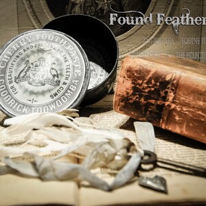 May include: A vintage tin container for Goodrick Toowoomba Cherry Toothpaste. The tin is silver with black lettering and a crest featuring a kangaroo and an emu. The tin is sitting on a book with a brown leather cover.