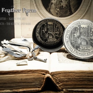 May include: Two vintage round tin boxes with black lids and white labels. The labels feature a black and white illustration of a building with the text "YOLD GLYSSCH GERRYE TOOTHPASTE 1746 MAYSTRES ENEWBERY & SONS. LONDON." The boxes are sitting on an open book with a key and some fabric scraps.