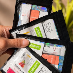 May include: Three black resealable pouches containing Herbalife Nutrition products. Each pouch features a clear window displaying individual packets of Herbal Tea Concentrate, Skin Collagen Beauty Booster, and Liftoff drink mix in various colors.