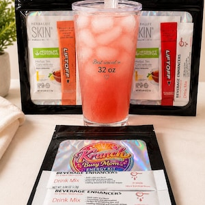 May include: A clear plastic tumbler filled with pink liquid and ice, featuring the text 'Best served in 32 oz' with a heart icon. It is surrounded by various Herbalife Skin and Krunch Busy Mom Energy Kit beverage enhancer packets and drink mix sticks.