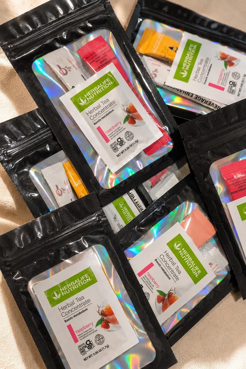 May include: Multiple black resealable pouches containing Herbalife Nutrition Herbal Tea Concentrate in raspberry flavor. Each pouch features a clear window displaying the product label with the text Herbal Tea Concentrate, raspberry, and 0.06 oz.