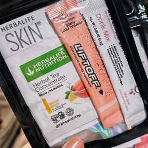 May include: A black pouch containing Herbalife Skin and Nutrition products. Visible items include a Herbal Tea Concentrate packet in lemon flavor, 0.06 oz, and a Liftoff effervescent energy supplement drink mix packet in tropical fruit flavor, 0.08 oz.