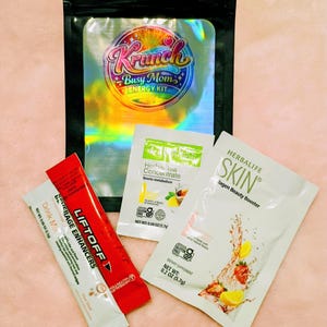 May include: A black holographic pouch labeled Krunch Busy Mom Energy Kit containing three individual packets: a red Liftoff beverage enhancer, a white Herbal Tea Concentrate packet, and a white Herbalife Skin Collagen Beauty Booster packet.