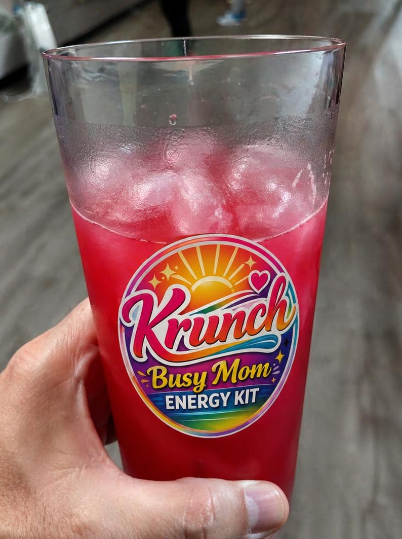 May include: A clear plastic tumbler filled with a bright pink liquid and ice cubes. A circular sticker on the front features a sun graphic, a heart, and the text Krunch Busy Mom ENERGY KIT in a colorful, rainbow-gradient font against a dark background.