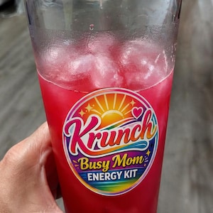 May include: A clear plastic tumbler filled with a bright pink liquid and ice cubes. A circular sticker on the front features a yellow sun, a small heart, and the text Krunch Busy Mom ENERGY KIT in colorful, stylized lettering against a rainbow background.