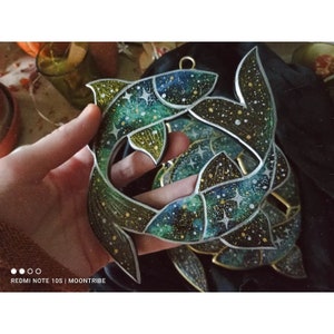 Op de afbeelding: Twee houten visornamenten geschilderd met een galaxy-ontwerp. De vissen zijn groen, blauw en zwart met gouden glitteraccenten. De vissen kijken elkaar aan en zijn met elkaar verbonden bij de staarten.