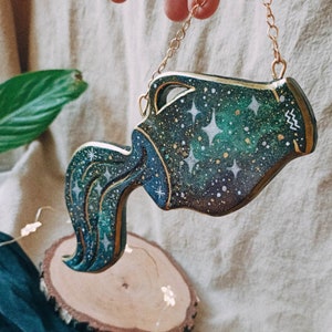 May include: A wooden cutout of the Aquarius zodiac sign, painted in shades of green and blue with gold accents. The cutout is shaped like a water pitcher pouring water, and it is decorated with white stars and a gold chain.