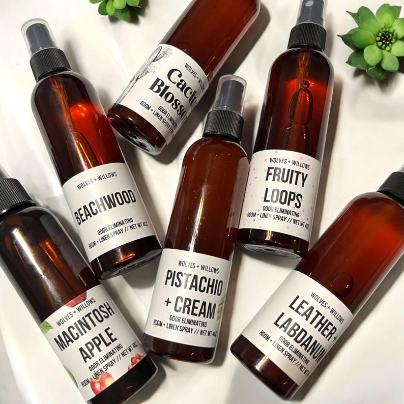 May include: A collection of ten amber glass spray bottles with white labels. The labels feature different scents, including Cactus Blossom, Beachwood, Macintosh Apple, Fruity Loops, Pistachio + Cream, and Leather Labdanum. Each bottle has the text "Wolves + Willows" and "Door Eliminating Room + Linen Spray" on the label.