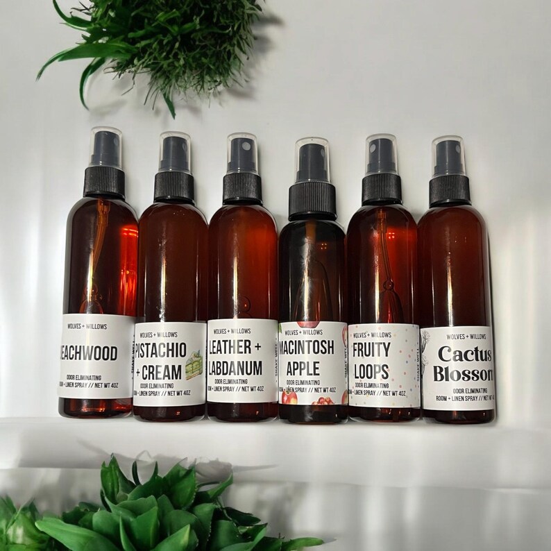 May include: Six brown glass spray bottles with white labels and different scents: Eachwood, Pistachio Cream, Leather + Labdanum, Macintosh Apple, Fruity Loops, and Cactus Blossom. The bottles are arranged in a row on a white surface.