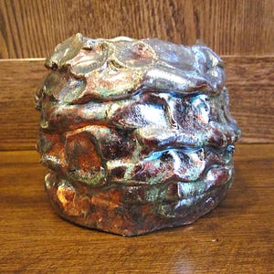 May include: A handmade ceramic pot with a textured, layered design. The piece features a blend of brown, bronze, and silver metallic glazes, with hints of green and blue. The unique shape and colour palette create a rustic, artistic aesthetic.