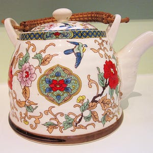 Könnte beinhalten: Eine Keramik-Teekanne mit weißem Grund, verziert mit Blumen- und Vogelmotiven in Rot, Rosa, Blau und Gelb. Sie hat einen geflochtenen Griff und einen passenden Deckel mit einem kleinen Knopf. Ausguss und Griffe sind weiß.