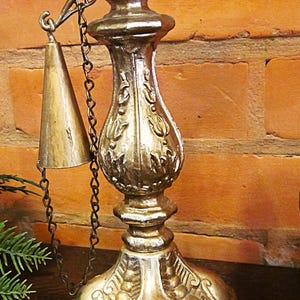 Peut inclure: Un chandelier argenté orné avec une cloche attachée par une chaîne. Le chandelier a une base large avec des volutes décoratives, une colonne centrale avec des détails en forme de feuilles et un dessus avec une plateforme circulaire. Fond de mur en briques.