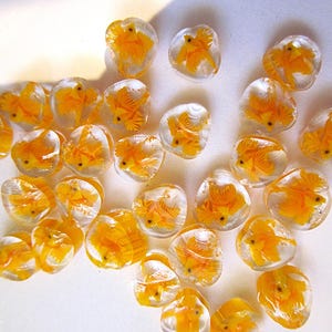 May include: A collection of clear, oval-shaped beads, each encapsulating a vibrant orange goldfish. The fish are detailed, with visible fins and tails. The beads are scattered across a white surface, creating a playful and aquatic theme.