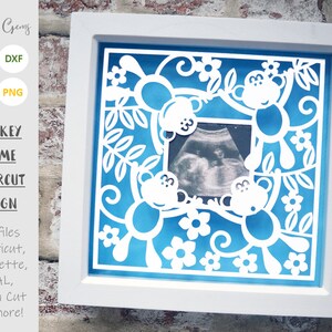 Può includere: Una cornice bianca con sfondo blu e un design di carta tagliata bianca di scimmie e fiori. La cornice ha un'apertura trasparente per una foto, e la foto mostra un'immagine di ecografia di un bambino.